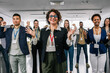 © Mediteraneo - Portrait of Hispanic woman CEO standing in front of her multiracial colleagues with their hands in the air, celebrating their success.