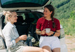 © Ilona - Two women friends are sitting in the back of car, talking and drinking coffee from a thermos during a trip out of city.