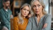 © Pinklife - An elderly woman with a worried expression has her arms crossed as a family interaction occurs in the blurred background, suggesting emotional tension or concern.