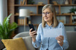 © Liubomir - A smiling senior woman sits on sofa, using smartphone and reading a document, in a cozy home setting.