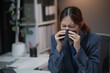 © amnaj - Woman in a blue suit sneezing into a tissue while sitting at her office desk, looking unwell and ill, implying a flu or cold.