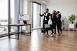 © sofiko14 - Business team in suits enjoying playful moment in office. Group of colleagues with woman on rolling chair showing teamwork and fun at work. Bright office space with large windows.