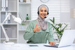 © Liubomir - Smiling customer service representative wearing a headset gives a thumbs up while working at her office desk with a laptop ,and lamp in a modern workspace.