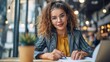 © Pinklife - A woman with curly hair and glasses is writing in a notebook. She is seated in a cozy cafe with modern decor and warm lighting, enjoying a drink beside her.