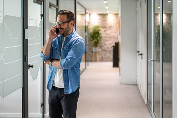 Wall Mural - Positive male financial advisor with glasses making phone calls while standing in office corridor