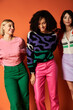 © LIGHTFIELD STUDIOS - Young, beautiful women in vibrant clothes stand together, showcasing friendship and cultural diversity on an orange background.