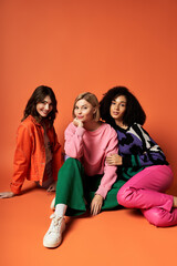 Wall Mural - Three young multicultural women in stylish outfits sitting closely together on an orange background.