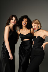 Wall Mural - Three young women, each wearing a black dress, standing gracefully together against a neutral grey backdrop.