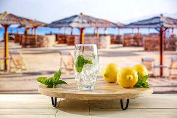 Sticker - A large glass of still mineral water with ice and citrus fruit on a wooden table against the background of a blue sky and a blurred beach. Vacation time and tropical beach