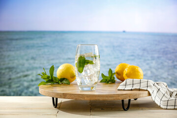 Sticker - Lemon fruits, fresh mineral water, ice in glass on wooden table top with beautiful blur landscape of calm blue sea and blue sky. Vacation time and cool drink for hot day.