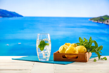 Sticker - Lemon fruits, fresh mineral water, ice in glass on wooden table top with beautiful blur landscape of calm blue sea and blue sky. Vacation time and cool drink for hot day.