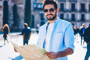 Half length portrait of happy bearded young man in black sunglasses smiling at camera while strolling in urban setting of old architectural city using map for searching showplaces and right direction