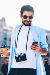 Wall Mural - Cheerful male tourist laughing during chatting online on smartphone and strolling with coffee to go in downtown during summer trip.Bearded traveler sharinf media in social networks walking outdoors