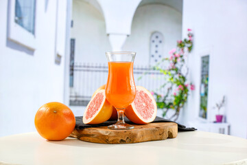 Wall Mural - Glass of citrus fruit juice and fruit on a wooden table against the backdrop of the blur white buildings. The sunlight and a summer background with space for food and drink and a product.