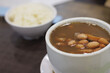© TimmyTimTim - A bowl of Singapore beans soup and rice is on a table. The soup is brown and has a lot of beans in it.