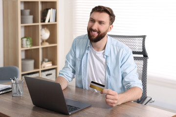 Wall Mural - Online banking. Happy young man with credit card and laptop paying purchase at table indoors