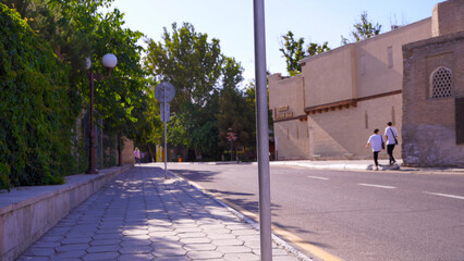 Wall Mural - A street with a sidewalk and a pole with a sign on it. Two people are walking on the sidewalk