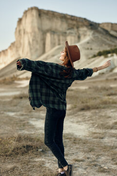 Outdoor adventure woman in plaid shirt and hat embracing nature with arms outstretched in front of mountain