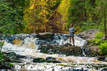 A woman stands on rocks by a rushing waterfall in the forest, enjoying the natural beauty and serene environment. The lush greenery and flowing water create a perfect scene for nature and adventure