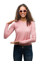 Sticker - Beautiful young hispanic woman wearing sunglasses gesturing with hands showing big and large size sign, measure symbol. Smiling looking at the camera. Measuring concept.