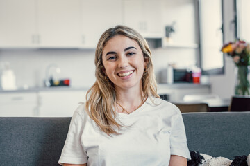 Wall Mural - Happy attractive young adult woman housewife sitting on sofa looking at camera in modern cozy home, smiling single 20s lady beautiful face relaxing on couch alone enjoy wellbeing posing for portrait.