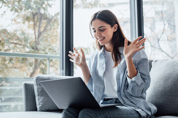 Smiling girl sit on couch at home wave talk with friend or family having video call on laptop, happy young woman greeting say hello, speak communicate using dating online application on computer.