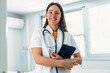 © nenetus - Confident female doctor using her digital tablet whilelooking at camera standing in the consultation.