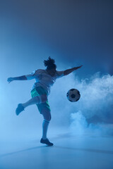 Wall Mural - A female athlete kicks a soccer ball in a blue smoky studio setting.