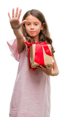 Wall Mural - Brunette hispanic girl holding a gift with open hand doing stop sign with serious and confident expression, defense gesture