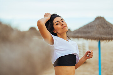 Wall Mural - A smiling young Hispanic female radiates happiness on a sandy beach, dressed in casual summerwear with a straw shelter in the background.