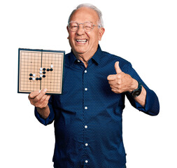 Sticker - Senior man with grey hair holding asian go game board smiling happy and positive, thumb up doing excellent and approval sign