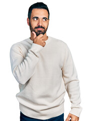 Poster - Young hispanic man with beard wearing casual white sweater with hand on chin thinking about question, pensive expression. smiling with thoughtful face. doubt concept.