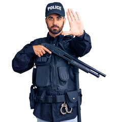 Canvas Print - Young hispanic man wearing police uniform holding shotgun with open hand doing stop sign with serious and confident expression, defense gesture