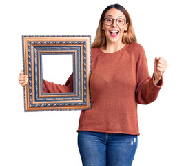 Sticker - Beautiful young woman holding empty frame screaming proud, celebrating victory and success very excited with raised arms