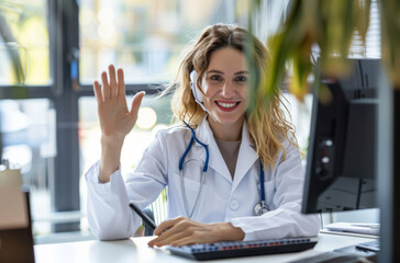 Wall Mural - A woman is sitting at her desk, waving to the doctor on screen who appears happy and relaxed in his medical gown with stethoscope around neck