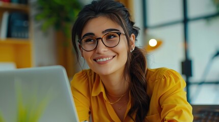 Aufkleber - A young woman with long dark hair and glasses smiles while working on a laptop computer. She is dressed in a yellow blouse and appears to be in a modern office setting.