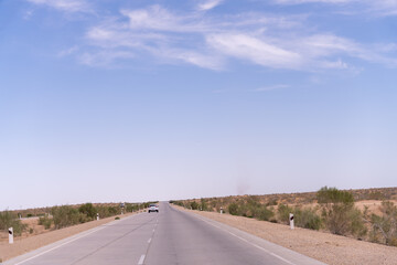 Wall Mural - A long, empty road with a car driving down it. The sky is clear and blue. The road is surrounded by a desert landscape