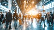 © vadosloginov - A busy trade show hallway filled with attendees walking in different directions, some stopping to talk or look at displays. The space is bright and modern, with high ceilings and large windows.