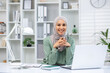 © Liubomir - Portrait of a young Muslim woman in a hijab sitting in an office at a desk with a laptop, hands folded together near her face, smiling and looking at the camera.