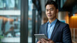 © ArtBox - Attractive young Chinese businessman using a digital tablet while standing in front of windows in office