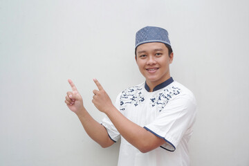 Muslim Asian man with happy expression pointing his finger to copy space. Ramadan and Eid Mubarak concept. Smiling Muslim Asian male pointing, isolated on white background.