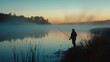 © stocker - A fisherman stands on the shore of a lake at dawn. The water is calm and still. The sky is a light blue and the sun is rising.