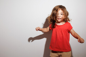 Cute brunette toddler girl with curly hair in red t-shirt smiling widely at white background 