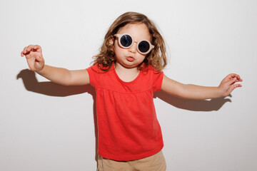 Cute brunette toddler girl in red t-shirt and round beige sunglasses having fun raising hands at the white background, kids face expressions 