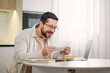 © Ivan - Smiling young man holding paper bill letter while working at table with portable laptop indoors. Skilled caucasian freelancer feeling shock while reading received information at home interior.