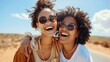 © ifoto - Two young women laughing and hugging on the side, wearing sunglasses in front of an open road. The woman is black with curly hair
