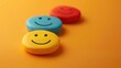 © Naret - Three colorful smiley face erasers on an orange background, representing happiness, positivity, and simplicity in a fun and playful setting.