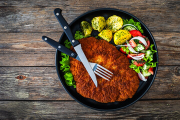 Poster - Crispy breaded fried pork chop, boiled potatoes and fresh vegetables served on black plate on wooden table
