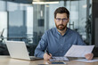 © Tetiana - Portrait of a young man in glasses and a blue shirt sitting in the office at a table with a laptop, engaged in data analysis and statistics, looking seriously at the camera
