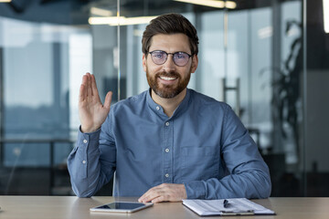 Wall Mural - Portrait of a smiling young man in a blue shirt and glasses sitting at a desk in the office, talking on a video call, greeting and waving at the camera